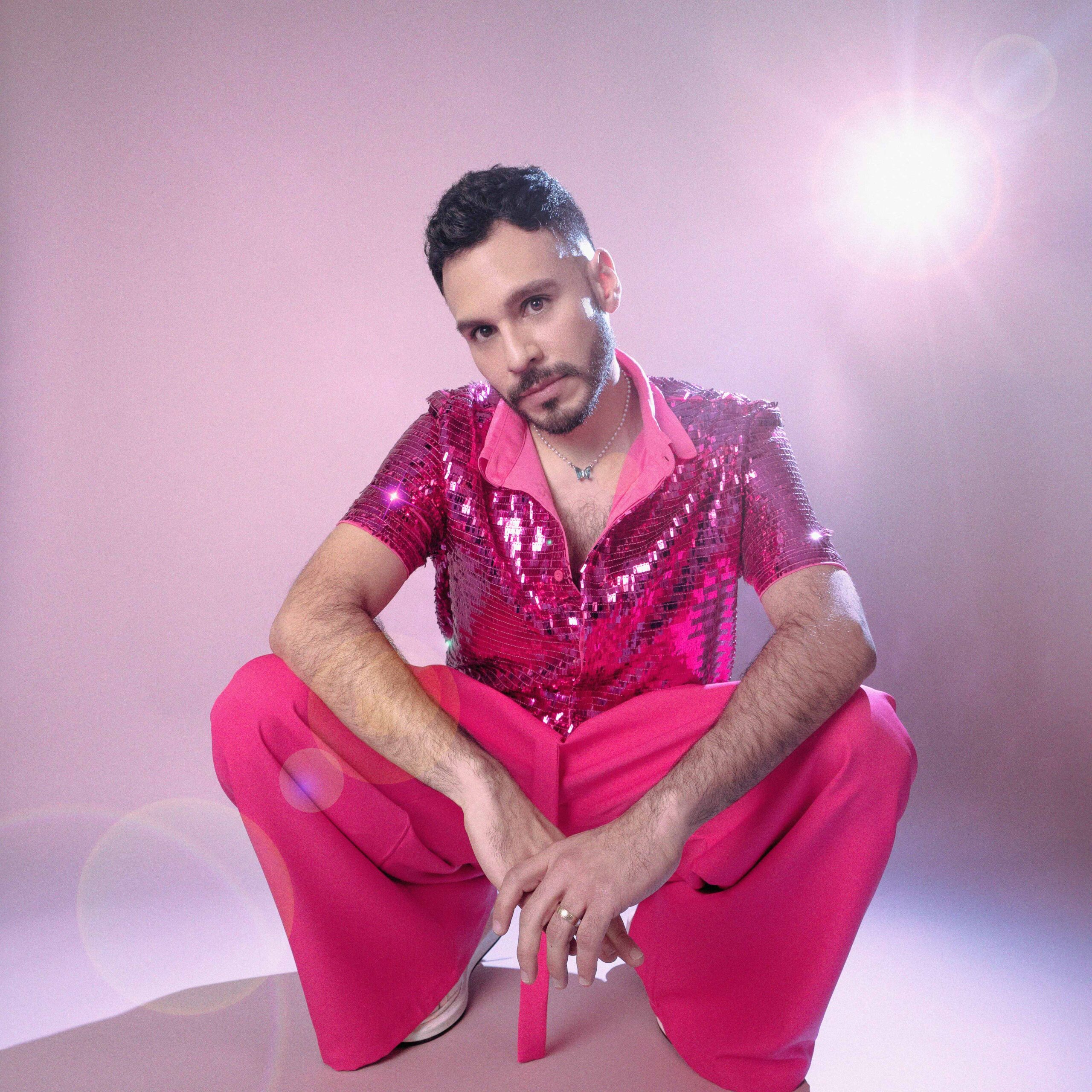 Fhernando sentado con pantalon oversize acampanado colro rosa y camisa con lentejuelas color rosa.